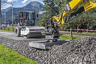 Ein gelber Bagger verdichtet frischen Schotter auf einer Strasse, während im Hintergrund eine weisse Walze bereitsteht. Rechts und links der Strecke sind Wiesen, Bäume und Schutzgeländer zu sehen. 