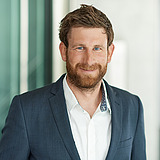 Technischer Bereichsleiter im Anzug lächelt in die Kamera. Im Hintergrund ist ein Lift zu sehen.