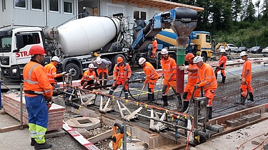 STRABAG - Mitarbeiter führen auf einer Baustelle in Lindau einen Testeinbau von Basaltfaserbeton durch; im Hintergrund stehen Betonmischer und Baumaschinen.
