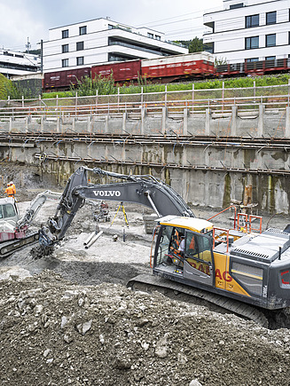 Überblick über die Baugrube: Zu sehen sind ein Bagger im Vordergrund beim Aushub der Grube. Im Hintergrund ist ein Mini-Bagger sowie ein weiteres Baugerät zur Spannung der Anker ersichtlich. 