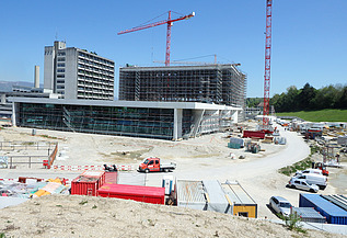 Grossbaustelle mit mehreren Gebäuden im Bau, darunter ein modernes, teilweise verglastes Gebäude im Vordergrund sowie ein grosses eingerüstetes Bauvolumen dahinter. Zwei rote Turmdrehkrane ragen in den Himmel. Auf dem Gelände befinden sich zahlreiche Baucontainer, Maschinen, Fahrzeuge und Baumaterialien. Rechts ein Baustellenweg, links Bauabsperrungen und Lagerflächen.