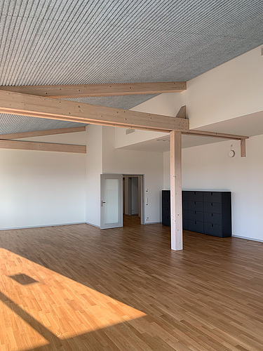 Heller, leerer Innenraum mit Holzparkett, sichtbaren Holzbalken und Akustikdecke, moderner Tür und schwarzen Schränken an der Wand.