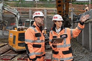 Frau mit Helm und Baustellenkleidung erklärt Mann die Baustelle