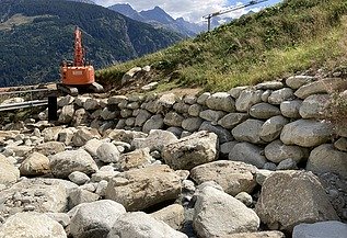 STRABAG-Bagger baut Steinschutzwand in den Bergen am Aclettbach bei Disentis. Im Hintergrund ist eine Berglandschaft zu sehen.
