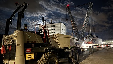 Ein STRABAG-Dumper steht nachts auf der Baustelle; im Hintergrund arbeitet ein grosser Raupenkran vor der Schaffhauserstrassen-Brücke.
