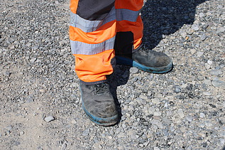 Bauarbeiter in orangefarbener Warnkleidung steht mit robusten Arbeitsschuhen auf einer kiesigen Fläche.