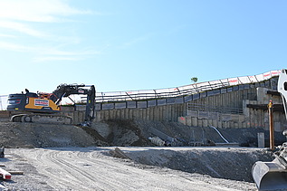 Gelber Bagger in einer Baugrube auf einer Anhöhe bei der Arbeit. Im Hintergrund ein strahlend blauer Himmel. 