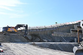 Gelber Bagger in einer Baugrube auf einer Anhöhe bei der Arbeit. Im Hintergrund ein strahlend blauer Himmel. 