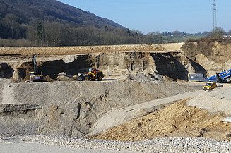 Weitläufige Kiesgrube mit Baggern und Lastwagen, die Sand und Kies abbauen, im Hintergrund sind Hügel und Bäume zu sehen.