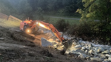 Arbeiten bei Dämmerung - STRABAG Ingenieurbau STRABAG Bagger arbeitet bei Abenddämmerung im Scheinwerferlicht am Aclettbach in Disentis. Im Hintergrund kann begrünte Bäume.