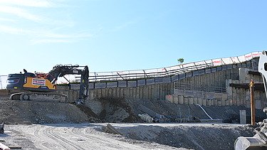 Gelber Bagger in einer Baugrube auf einer Anhöhe bei der Arbeit. Im Hintergrund ein strahlend blauer Himmel. 