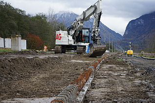 Ein Bagger arbeitet auf einer Erdbaustelle und zieht Material entlang. Im Vordergrund liegen eng aneinandergereihte Armierungseisen, im Hintergrund sind Bäume und eine Bergkulisse zu sehen.