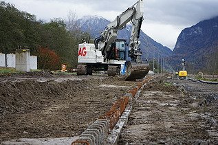 Ein Bagger arbeitet auf einer Erdbaustelle und zieht Material entlang. Im Vordergrund liegen eng aneinandergereihte Armierungseisen, im Hintergrund sind Bäume und eine Bergkulisse zu sehen.