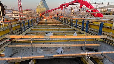  Ein Kran der STRABAG hebt nachts ein langes Brückenteil über Bahngleise. Die Baustelle ist hell erleuchtet.