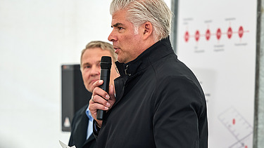 Ein Redner in dunkler Jacke spricht bei einer Veranstaltung in Bürglen in ein Mikrofon. Hinter ihm steht eine weitere Person. Im Hintergrund ist ein Banner der Veranstaltung zu sehen.