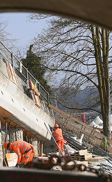 Mehrere STRABAG-Arbeiter in orangefarbener Schutzkleidung sind im Hintergrund an einer Betonwand tätig.