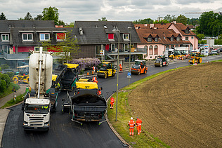 Baumaschinen und Arbeiter tragen frischen Asphalt auf eine Strasse in einem Wohngebiet auf.