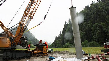 Baustellenkran hebt Filterbrunnen hoch, während STRABAG Bauarbeiter am Ziel warten. Im Hintergrund sind grüne, bewaldete Berge zu sehen.