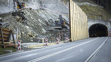 Mehrere Bagger in einer Baugrube am Fels abspitzen, rechts ist der Sisikoner Tunnel zu sehen