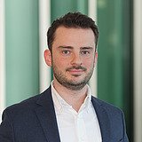 Technischer Bereichsleiter im Anzug lächelt in die Kamera. Im Hintergrund ist ein Lift zu sehen.