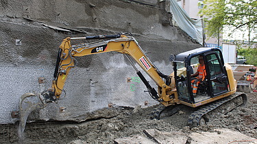 Ein gelber Bagger mit STRABAG-Logo arbeitet in einem leicht abschüssigen Graben an einer Betonwand. Ein Bauarbeiter sitzt in der Fahrerkabine.