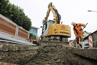 Liebherr-Bagger bei Aushubarbeiten in einer offenen Baugrube. Ein Bauarbeiter in orangefarbener Schutzkleidung säubert Grabenrand mit einem Besen. Im Hintergrund sind Werkleitungen gestapelt. Absperrungen und Verkehrsschilder sichern den Baustellenbereich.