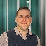 Kommunikationsleiter lächelt in die Kamera. Im Hintergrund ist ein Lift zu sehen.