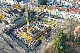 Luftaufnahme einer Baustelle mit Baugrube und Kränen in einem Stadtgebiet.