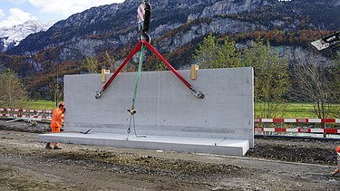 Eine schwere Betonelementwand hängt an roten und grünen Hebegurten unter dem Kranhaken. Auf der Baustelle stehen Mitarbeitende in orangefarbener Schutzausrüstung daneben, während im Hintergrund eine Berglandschaft mit bewaldeten Hängen zu sehen ist.