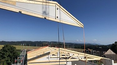 Rohbau der Labor-Erweiterung mit dem Einsatz eines Krans für eine weitere weisse vorgefertigte Wand und sichtbaren Holzbalken für das Dach. Im Hintergrund ländliche Umgebung mit Feldern, Bäumen und Hügeln.