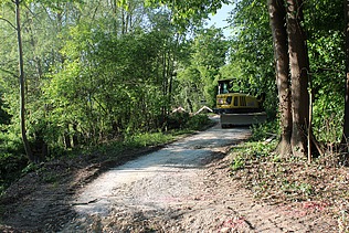 Ein gelber Bagger von STRAGAG steht auf einem geschotterten Waldweg, umgeben von dichten grünen Bäumen und Sträuchern.