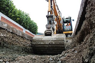 Bagger beim Aushub einer Baugrube, aufgenommen aus der Perspektive der Grubensohle. Der Baggerführer sitzt in der Kabine und steuert die Schaufel, die gerade Erdmaterial bewegt.