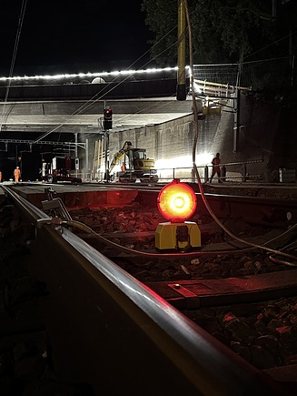 Scheinwerfer und Warnleuchten beleuchten die Gleise, während Arbeiter im Hintergrund an einer provisorisch eingerüsteten Konstruktion arbeiten.