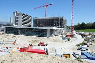 Grossbaustelle mit mehreren Gebäuden im Bau, darunter ein modernes, teilweise verglastes Gebäude im Vordergrund sowie ein grosses eingerüstetes Bauvolumen dahinter. Zwei rote Turmdrehkrane ragen in den Himmel. Auf dem Gelände befinden sich zahlreiche Baucontainer, Maschinen, Fahrzeuge und Baumaterialien. Rechts ein Baustellenweg, links Bauabsperrungen und Lagerflächen.