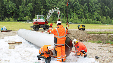 Drei STRABAG Bauarbeiter in orangefarbener Arbeitskleidung bereiten den Filterbrunnen für das Einsetzen mit einem Kran vor; im Hintergrund ein Bagger und Bäume.