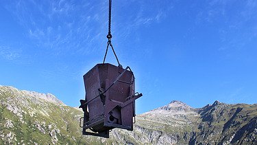 Ein Kübel für Beton wird vor dem blauen Himmel und einer Bergkulisse in der Schweiz über die STRABAG Baustelle gehoben.