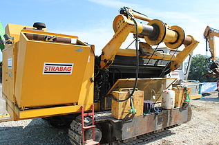 Gelbe Baumaschine mit mehreren Walzen und Anbaugeräten steht auf einer Baustelle im Hintergrund sind weitere Maschinen und Material zu sehen.