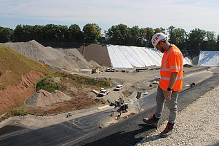 Bauarbeiter in orangefarbener Schutzkleidung steht am Rand einer steilen Böschung und blickt hinunter auf die Baustelle.