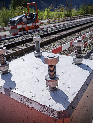 Ein Betonsockel steht direkt neben den Bahngleisen. Die Sonne wirft harte Schatten auf die Oberfläche, und im Hintergrund sind weitere Gleise und Absperrungen entlang der Strecke zu sehen.