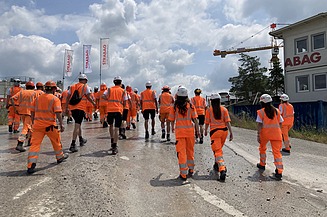 Eine Gruppe von STRABAG - Lernende in orangefarbener Arbeitskleidung und weissen Helmen geht auf eine Baustelle.