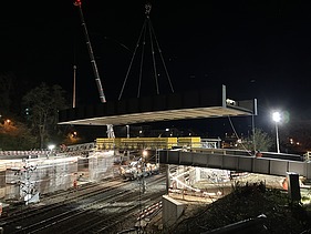 Bei Nacht wird ein grosses Brückenelement mit zwei Pneukranen über die Bahnlinie gehoben. Unterhalb der schwebenden Brücke sind die beleuchteten Gleise und Baustellenbereiche zu sehen. Mehrere Arbeiter sind auf der Baustelle im Einsatz.