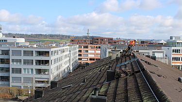 STRABAG Sicherheitsbeauftragter auf einem Dach mit Sicherungsseil. Im Hintergrund weitere Wohnblocks und eine blauer Himmer.