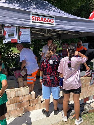 STRABAG-Zelt: Kinder und Erwachsene bauen unter Anleitung eine Mauer aus Ziegeln.