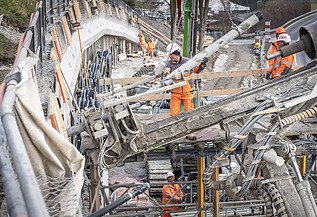 Nahaufnahme eines Bohrgeräts auf einer Baustelle. Mehrere STRABAG-Arbeiter in orangefarbener Schutzkleidung sind im Hintergrund an einer Betonwand tätig.