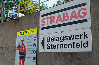 Betonmauer entlang einer Strasse mit einem grossen STRABAG-Schild und der Aufschrift ‚Belagswerk Sternenfeld‘, daneben ein Warnschild.