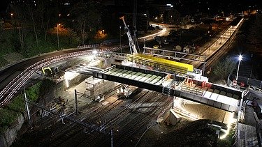 Baustelle in Küssnacht bei Nacht, beleuchtet von Scheinwerfern. Über den Bahngleisen wird eine neue Bahnbrücke mit einem grossen Kran eingesetzt. Im Hintergrund sind ein Bahnhof, Strassen mit Lichtspuren und beleuchtete Gebäude zu sehen.
