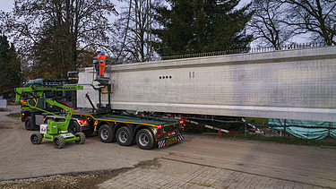 Ein grosser Turnhallenträger liegt auf einem langen Transportfahrzeug auf einer Strasse.