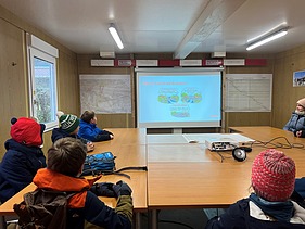 Eine Schulklasse sitzt in einem Baucontainer und verfolgt eine Präsentation zum Brückenbau auf einer Leinwand.