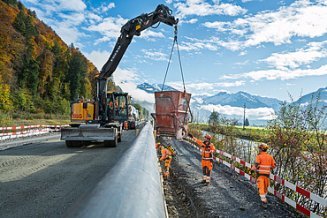 Ein Bagger hebt einen grossen Betonbehälter über eine Strassenbaustelle mehrere Arbeiter in oranger Schutzkleidung stehen daneben und sichern die Arbeiten. Im Hintergrund sind Herbstbäume zu sehen.