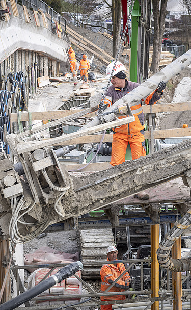 Baurabeiten am Hang - STRABAG Ingenieurbau Nahaufnahme eines Bohrgeräts auf einer Baustelle. Mehrere STRABAG-Arbeiter in orangefarbener Schutzkleidung sind im Hintergrund an einer Betonwand tätig.